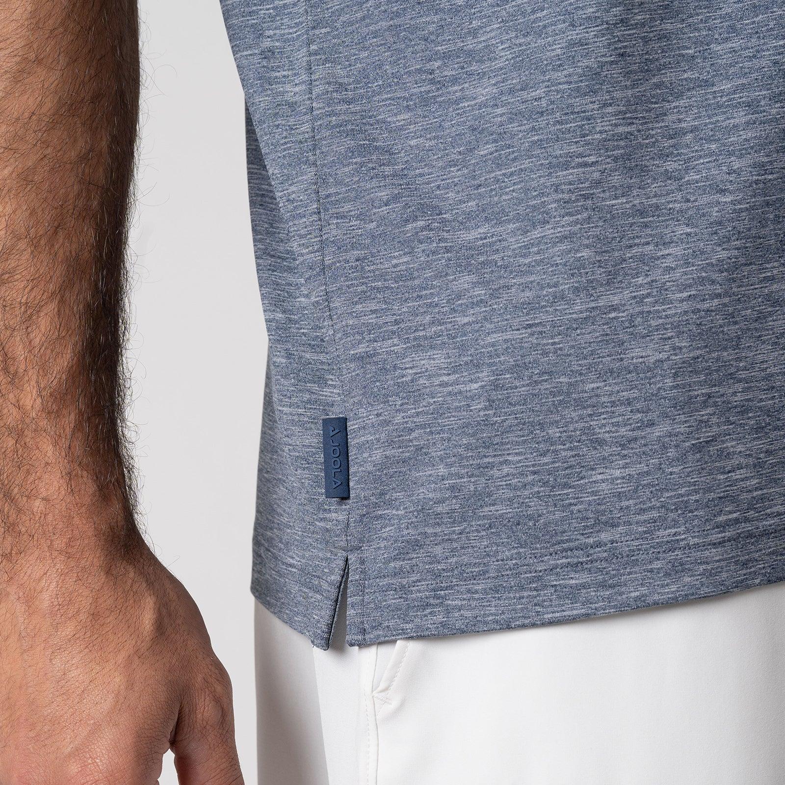 Nahaufnahme eines blaugrauen T-Shirts mit seitlichem Schlitz und kleinem Markenlabel am Saum, getragen von einer Person mit hellem Unterteil.