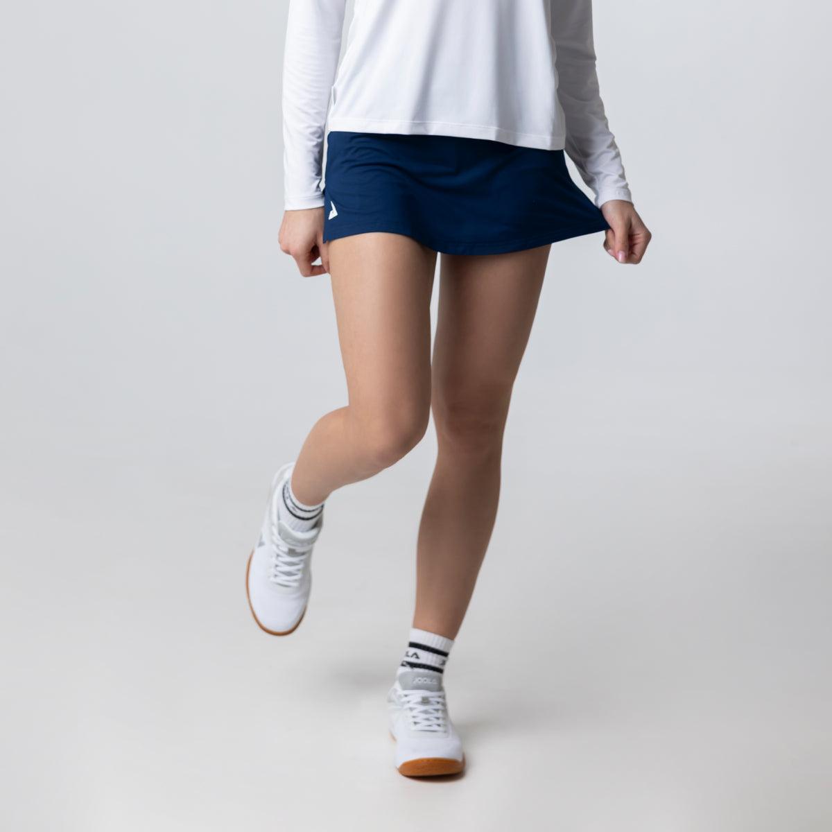 Frau trägt dunkelblauen Sport-Rock mit weißem Langarmshirt und weißen Sportschuhen.